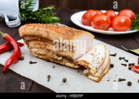 Un grande pezzo di pancetta cotta in forno con le spezie e le erbe su una carta con pomodori marinati. Un pasto di festa. Pasqua. Natale. Foto Stock