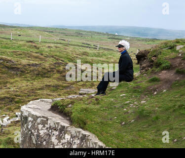 Senior donna contemplativa in nero seduto vicino a scogliere di Moher Irlanda su un ventoso pomeriggio con le mucche e il Blue Hills in background Foto Stock