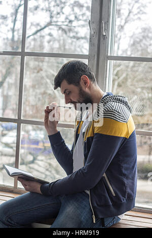 Ritratto di un informale barbuto hipster Uomo di pensiero e di rendere note in copybook o blocco note a casa. Grandi finestre backdround. scrittura a mano Foto Stock