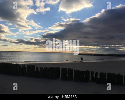 Persona cammina da sola lungo la spiaggia di sabbia al tramonto. Drammatica sweep di nuvole in cielo. Resti di un molo in legno, una fila di weathered posti di legno l Foto Stock