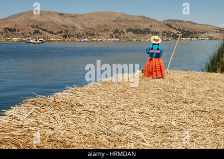 Una Donna vestita di tradizionale Peruviano abbigliamento guarda al lago Titicaca e la città di Puno dalle Isole Uros (o isole galleggianti) in Perù Foto Stock