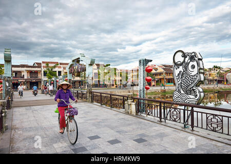 Una donna vietnamita che indossa un tradizionale cappello conico cavalca in bicicletta sul ponte Cau An Hoi nella città antica di Hoi An, provincia di Quang Nam, Vietnam. Foto Stock