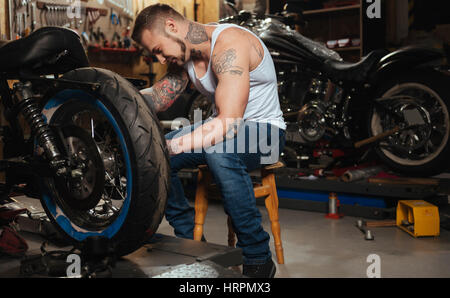 Solo un momento. Giovane uomo barbuto mantenendo il suo collo piegato, seduta in officina mentre si lavora con la sua moto Foto Stock