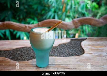 Bella di vetro turchese con fresco frullato di banana e un tubo di bambù su una trama di legno Scrivania con uno sfondo di vegetazione tropicale Foto Stock