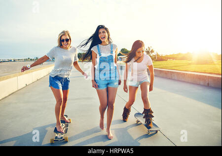 Tre active piuttosto giovane donna su una giornata al mare godendo le loro vacanze estive lo skateboard lungo una passeggiata lungomare a sunrise di ridere Foto Stock