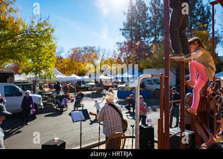 I bambini salire e giocare su una scala mentre un musicista bluegrass riproduce la musica durante un mercato agricolo in Danville, California, 3 dicembre 2016. Foto Stock