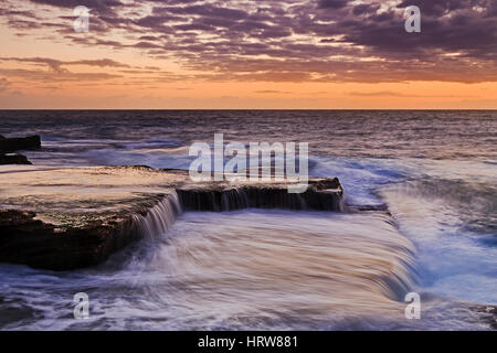 Colorato sunrise caldo da Maroubra Beach a Sydney. Navigare in flusso di onde piane su rocce di arenaria della costa australiana. Foto Stock