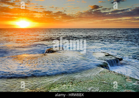 Luminoso colorato tramonto sull orizzonte a Maroubra Beach a Sydney. Onda in entrata fluisce sul piatto rocce costiere e alga verde. Foto Stock