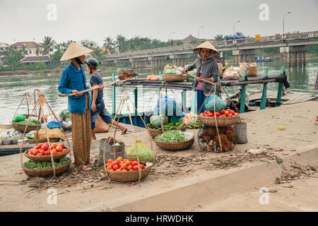 Hoi An, Vietnam - Marzo 14, 2014: Le donne nel cappello conico e l'uomo lo scarico di barca con merci per la vendita sul mercato dei prodotti freschi Foto Stock