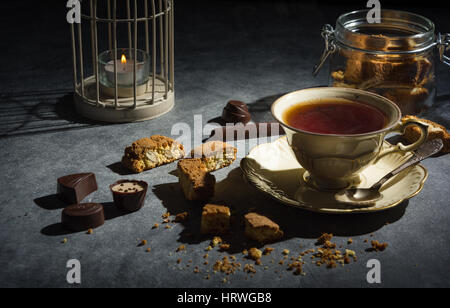 Tazza di tè , gustosi e sani biscotti di mandorle, ricco di vitamine e minerali in un vasetto di vetro e cioccolatini su uno sfondo scuro. Tasto basso. Foto Stock