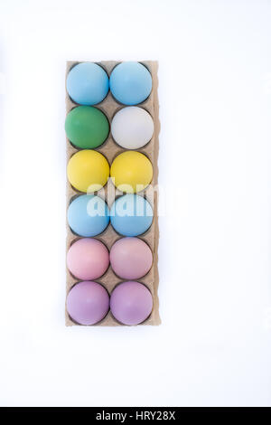 Uno bianco di un uovo di gallina e undici le uova di Pasqua di lilla tinti, rosa, blu, verde e giallo in una scatola di cartone da sopra con sfondo bianco, spazio di copia Foto Stock
