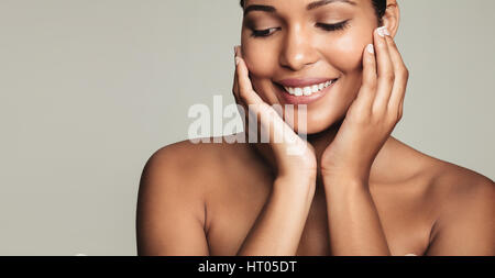 Primo piano della sorridente giovane donna con pulito e pelle sana su sfondo grigio. Bella femmina modello toccando il suo volto. Foto Stock