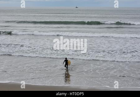 Un uomo in una muta, in possesso di una tavola da surf, passeggiate nel surf. Foto Stock