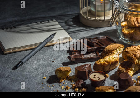 Gustosi e sani biscotti di mandorle, ricco di vitamine e minerali in un vasetto di vetro, cioccolatini e notebook su uno sfondo scuro. Tasto basso. Foto Stock