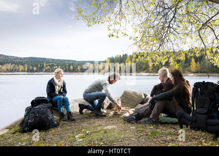 Amici guardando all uomo la preparazione per il falò sul lago campeggio durante l'autunno Foto Stock