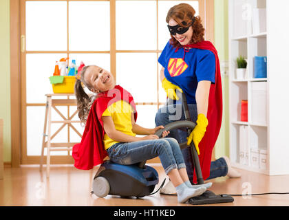 Superheros kid ragazza e la sua madre che fa servizio lavanderia insieme nel soggiorno. Famiglia - di mezza età della donna e bambino figlia hanno un divertimento mentre aspirapolvere Foto Stock