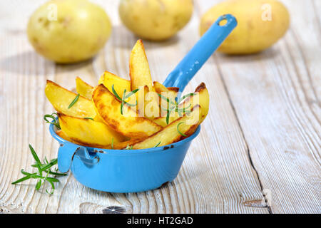 Patate al forno cunei con rosmarino servita in un vecchio malandato pot di smalto su una tavola di legno con tre patate in background Foto Stock