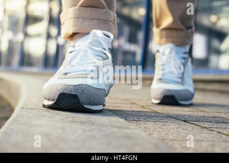 Gambe in pantaloni beige e bianco sneakers sono sul marciapiede. Close-up, angolo basso tiro, profondità di campo. Foto Stock