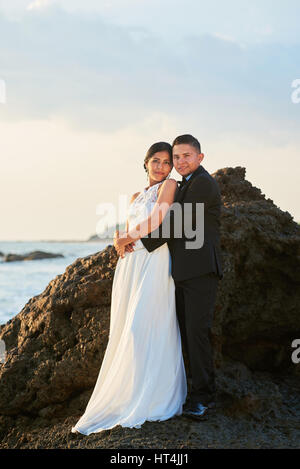 Ispanico sposa e lo sposo in piedi sulla roccia costa al tramonto luce arancione. Latino matura in amore giorno di nozze Foto Stock