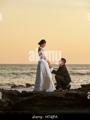 Lo sposo stand sul ginocchio e tenere premuto sposa mano sull'arancione tramonto sfondo nel mare spiaggia Foto Stock