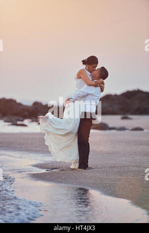 Felice baciare lo sposo e la sposa stand sulla fascia costiera sull'oceano nella luce del tramonto. Matrimonio esotico sulla spiaggia isola con rosa sullo sfondo del cielo Foto Stock