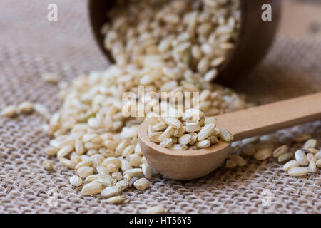 Eventuali fuoriuscite di grani di riso marrone sparsi su un letto di sacco, ciotola di legno e cucchiaio, vicino, stile rustico Foto Stock