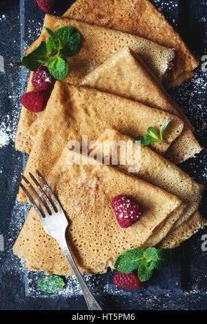 Deliziosa casa di grano intero crepes servito con lamponi freschi, foglia di menta e zucchero in polvere in ardesia. Vista superiore, closeup, immagine verticale Foto Stock