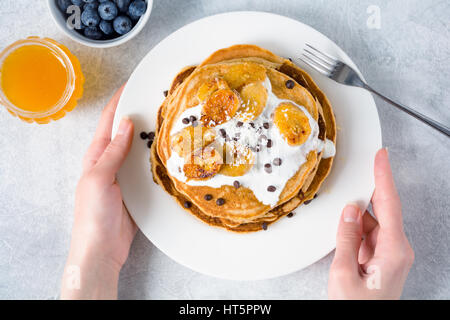 Pancake caramellato con banane, yogurt e scaglie di cioccolato sulla piastra bianca. Ragazza mangiare pancake per colazione. Prospettiva in prima persona, vista dall'alto Foto Stock