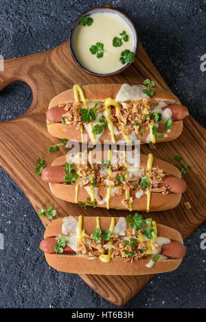 Hot Dog con salsiccia, cipolla fritti, foglie di coriandolo, salsa al formaggio e senape, servita su legno che serve oltre il bordo nero texture concreto sfondo. Foto Stock