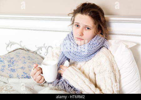 Malata con tazza di tè. Primo piano immagine di giovani frustrati donna in maglia Sciarpa blu tenendo una tazza di tè mentre è seduto nel letto della sua stanza. looki Foto Stock