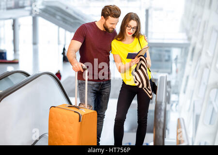 Giovane coppia felice di alzarsi con i bagagli su per la scala mobile per la zona partenze dell'aeroporto durante le loro vacanze estive Foto Stock