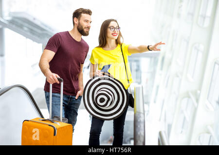 Giovane coppia felice di alzarsi con i bagagli su per la scala mobile per la zona partenze dell'aeroporto durante le loro vacanze estive Foto Stock