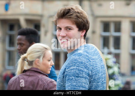 Un felice studente spire di sorridere alla telecamera come fa il suo modo per le sue lezioni. Egli sta portando il suo sacco sulle spalle e i suoi amici sono in b Foto Stock