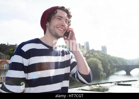 Un felice giovane sorride come utilizza il suo telefono cellulare. Egli è su un ponte su un fiume con la cattedrale in background. Foto Stock