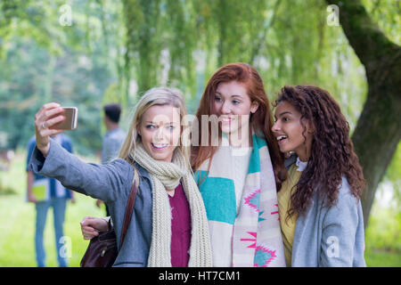 Tre femmine felice sorriso che prendono un selfie all'esterno. Essi sono avvolti fino a caldo come stanno sotto un albero. Foto Stock