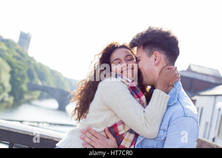 Una felice coppia giovane sorriso come rilassarsi all'aperto lungo il fiume. Essi sono avvolti fino a caldo per l'inverno in un abbraccio. Foto Stock