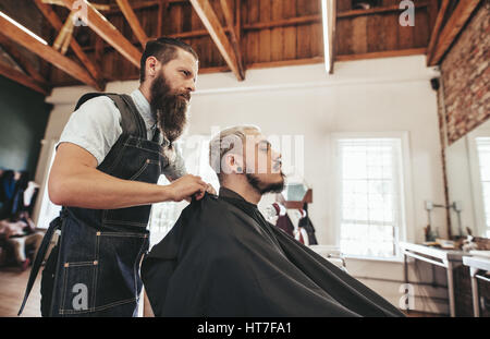 Vista laterale colpo di giovane uomo getting haircut in barberia. Barbiere di servire client nel salone di parrucchiere. Foto Stock