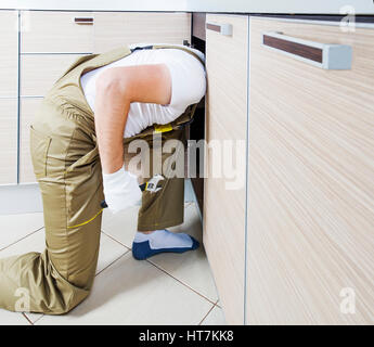 Plumber fissaggio di tubazioni in cucina. Plumber testa sotto lavello. Lavoratore con chiave di riparazione tubazioni fognature. Impianto idraulico funziona sullo sfondo. Foto Stock