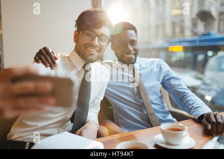 Giovani attraenti barbuto manager in bicchieri e il suo sorriso bello collega afro-americano che posano per una selfie pur avendo la pausa pranzo al cafe'. Foto Stock