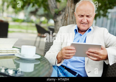 Ritratto di uomo anziano tenta di utilizzare il tablet, guardando curioso e confuso, mentre vi rilassate presso area salotto all'aperto del cafe sul giorno di estate Foto Stock