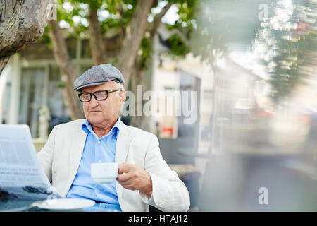 Ritratto di tipico vecchio uomo che indossa il cappuccio e cerchio nero, occhiali da lettura quotidiano fresco e avente tazza di caffè a outdoor cafe su mattinata estiva Foto Stock