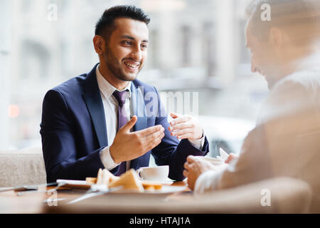 Ritratto di giovane entusiasta del Medio Oriente gestualità uomo attivamente e sorridente a parlare con il suo partner commerciale o di un collega in una tranquilla zona calda accesa cafe o Foto Stock