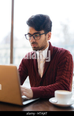Ritratto di giovane occupato medio orientali imprenditore indossando Felpa casual e cerchio nero occhiali seduto contro il vetro lavorando al computer portatile, cercando inten Foto Stock