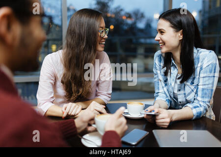 Il gruppo internazionale di giovani seduti a tavola rotonda in cafe contro la finestra con vista di sera le strade delle città, concentrarsi su due belle donne ridere, Foto Stock