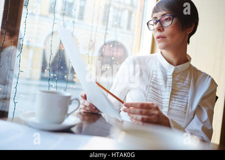 Ritratto di grave elegante imprenditrice che indossa una camicia bianca e occhiali lettura contratti e documenti mentre è seduto in cafe in corrispondenza della finestra tabella in EVA Foto Stock