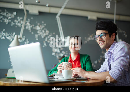 Ritratto di due giovani colleghi creativi l uomo e la donna sia di indossare un abbigliamento informale e bicchieri, lavorando alla riunione in cafe, sorridente e guardando l Foto Stock