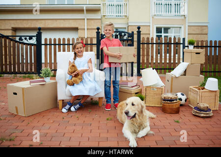 Ritratto di ordinata dei bambini felici, un ragazzo e una ragazza, ridendo e divertirsi mentre aspetta con scatole di cartone, i beni e i loro animali golden retriever o Foto Stock