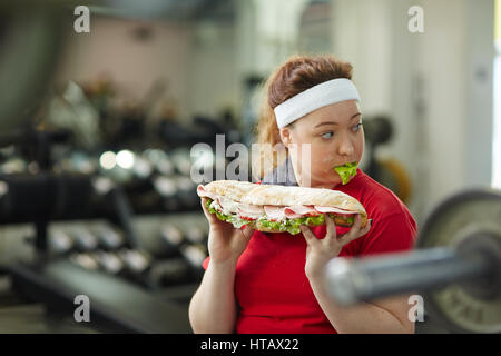 Ritratto di giovane donna sovrappeso mangiare grande unto ingrasso sandwich per allenarsi in palestra, concetto di cibo ossessione Foto Stock