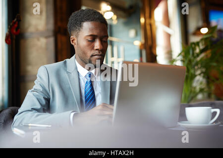 Ritratto di bello African- impresario americano indossare tuta formale incentrato guardando lo schermo del computer mentre occupato a lavorare con il computer portatile in cafe Foto Stock
