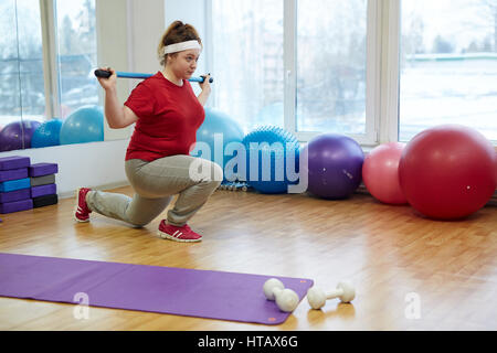 Vista laterale ritratto di determinata sovrappeso donna che lavorano in studio fitness: facendo split squat con corpo bar circondato con attrezzature per il fitness Foto Stock